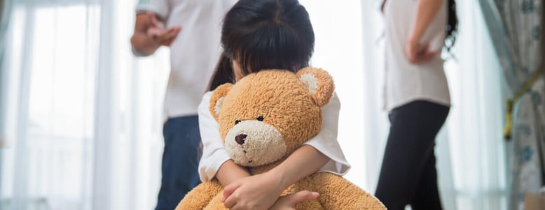 child hugs teddy bear while parents argue