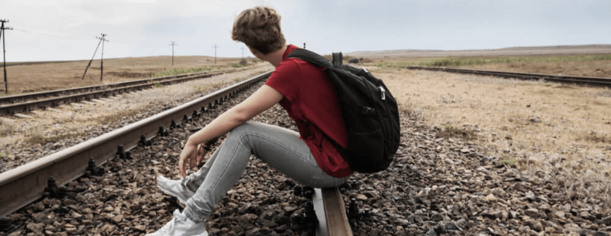 Teen boy skipping school, sitting on railroad tracks with backpack on